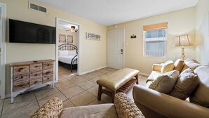 A living room with a beige sofa, a wooden bench, a television, and a door leading to a bedroom. - ✨Beachfront Complex + Community Pool | Sleeps 4 | Sunrise Village 111✨ (Gulf Shores)