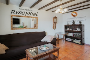 A living room with a dark brown sofa, a glass coffee table, a wooden bookshelf, a wall-mounted TV, and a decorative wall with text and a ring.