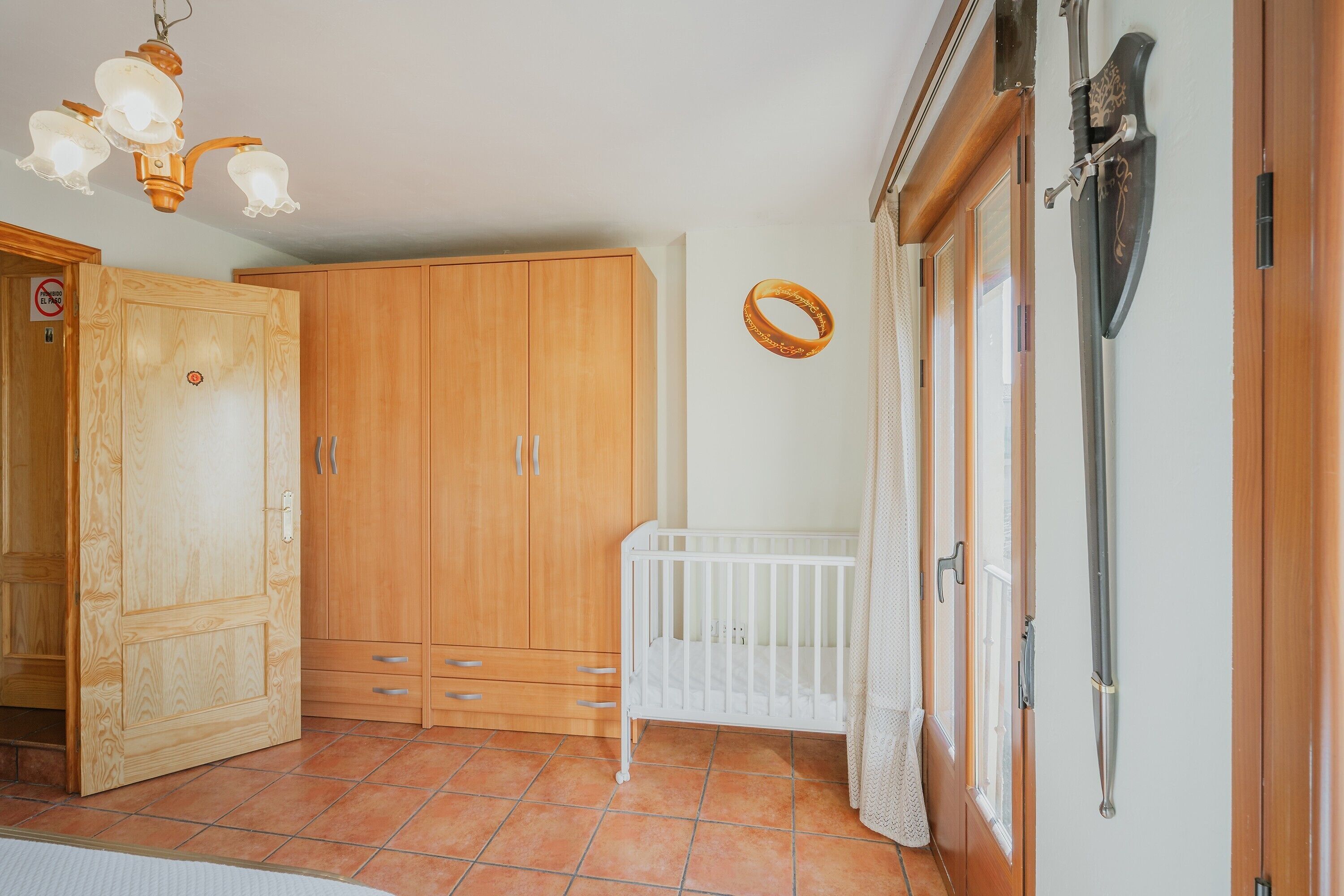A room with a wooden wardrobe, a white crib, a hanging sword, and a chandelier.