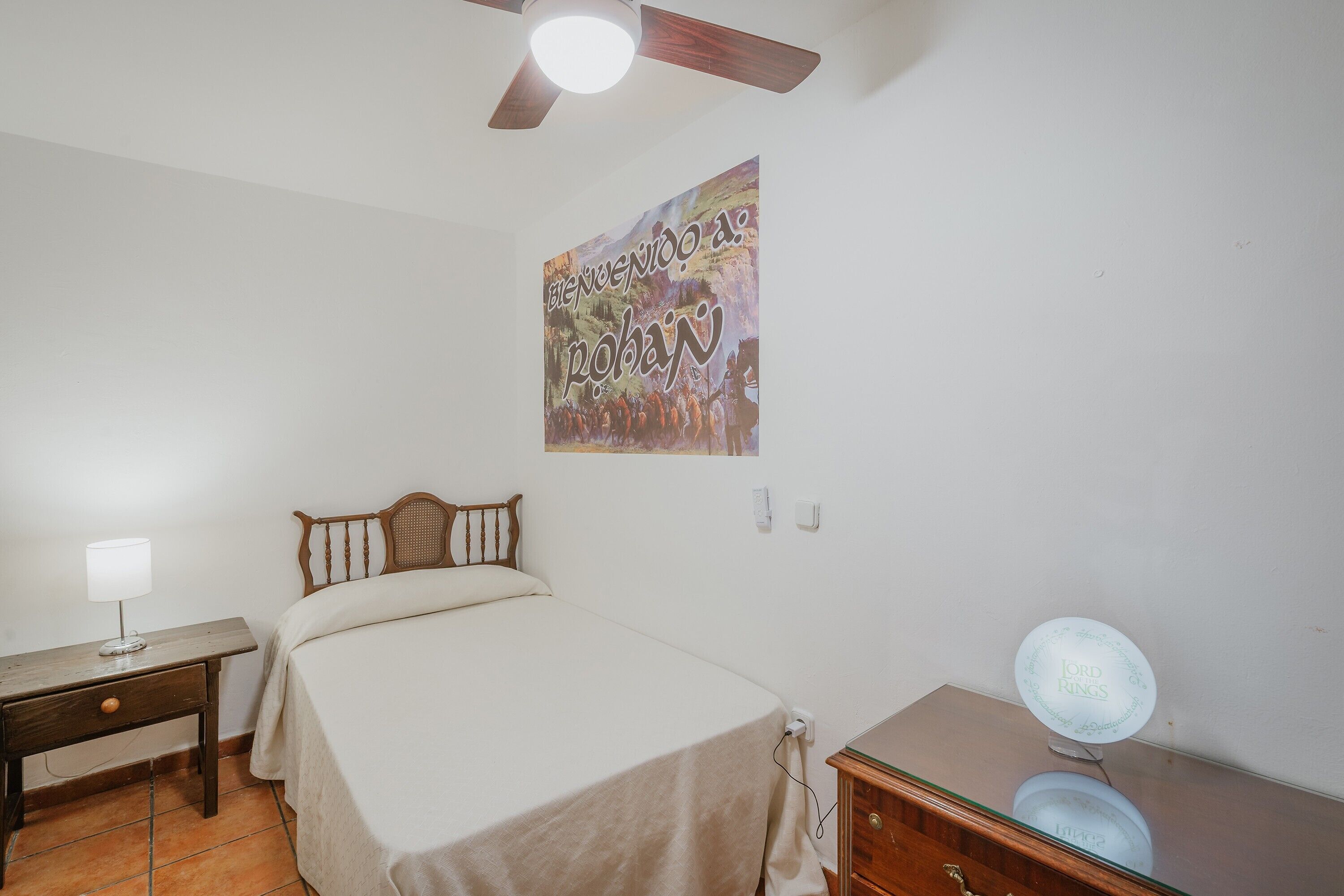 A bedroom with a bed, a desk with a lamp, a ceiling fan, and a poster on the wall.
