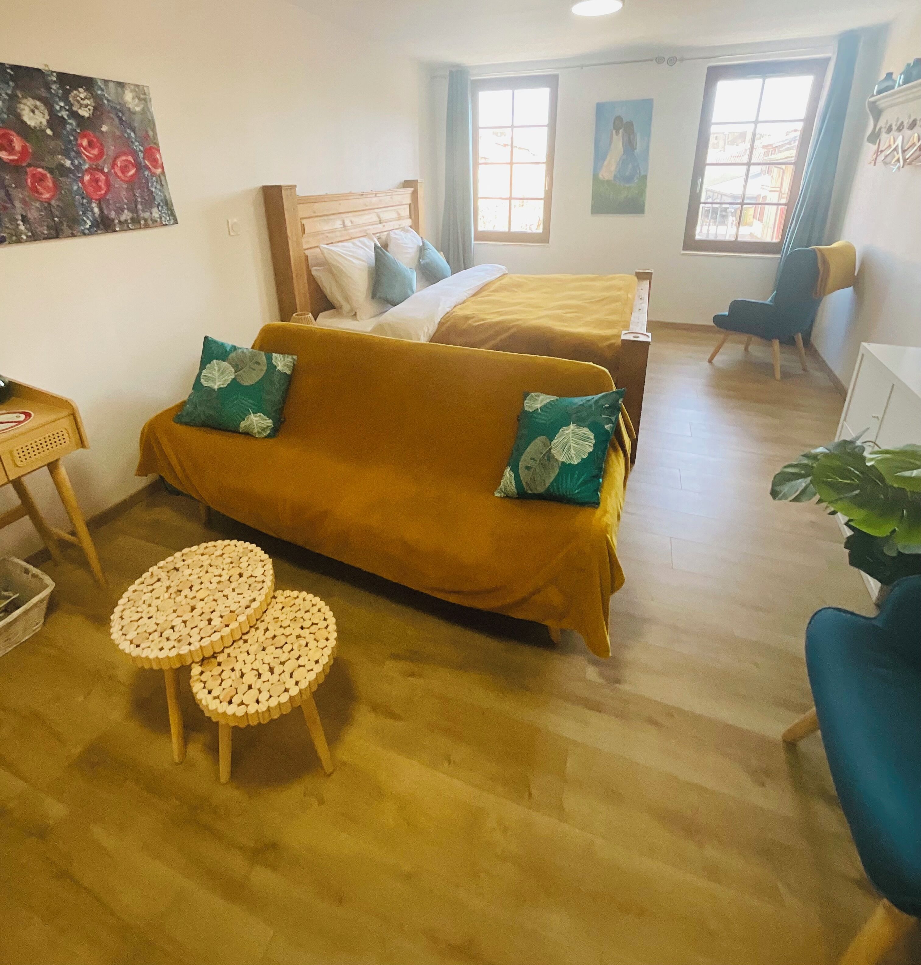 A cozy room with a sofa, two small round tables, a bed with a yellow bedspread, and two windows with curtains.