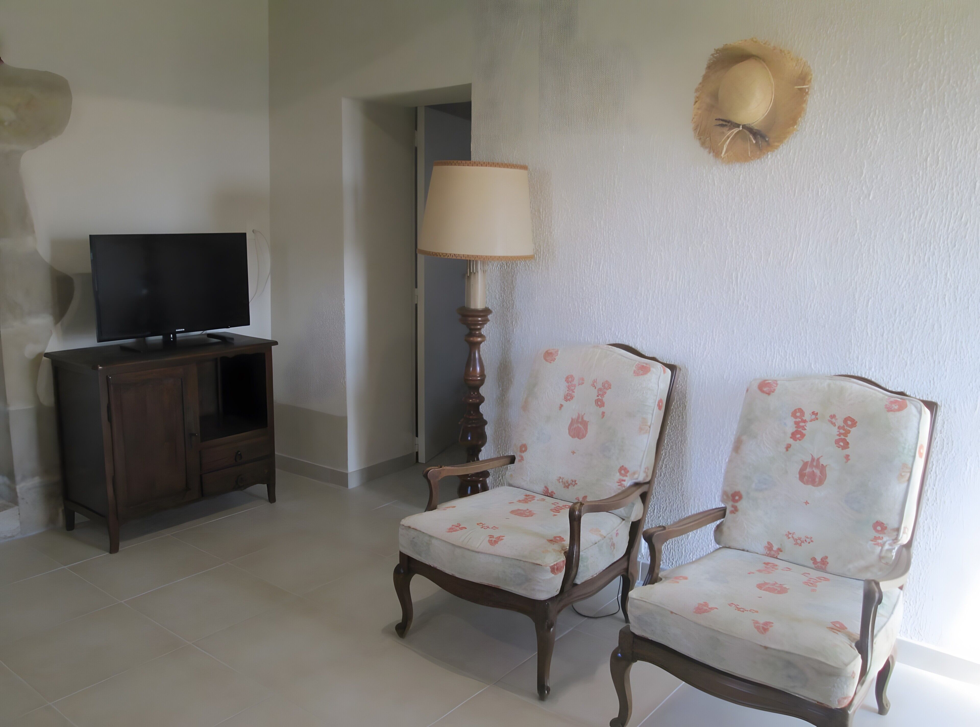 A room with a television on a wooden cabinet, two upholstered chairs with floral upholstery, a tall lamp, and a wall-mounted decorative item.