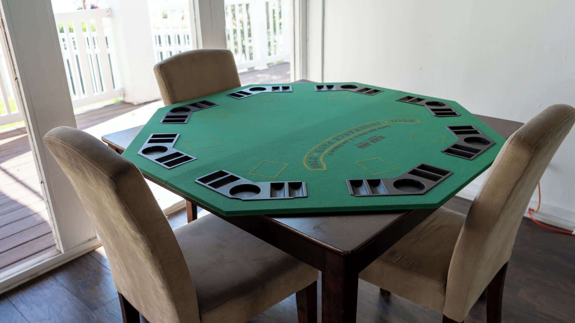 A green poker table with eight chairs, set on a wooden deck with a view of a white fence and buildings.