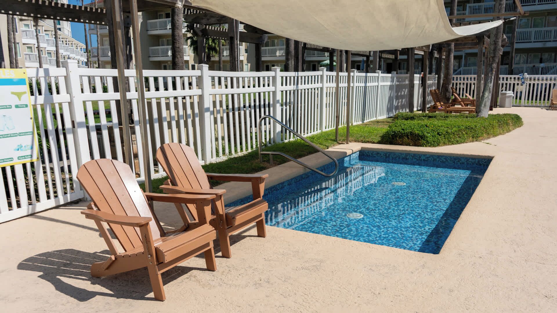 Un área de piscina con dos sillas Adirondack de madera, una valla blanca y un toldo con sombra.