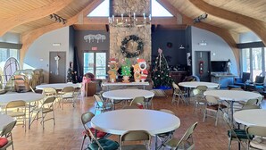 Ein großzügiger Raum mit hohen Holzbalken, einer großen Kristallleuchtre und einer Steinwand mit einem Weihnachtskranz. Tische und Stühle sind für Zusammenkünfte bereitgestellt.