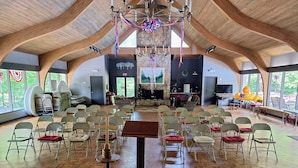 Ein geräumiger Saal mit hoher Holzbalkendecke, angeordneten Sitzplätzen, einem zentralen Tisch und einer Steinwand mit Kamin.