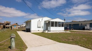 A single-story building with a covered porch, a concrete pathway leading to it, and a grassy area in front. - The Way Back: Cozy Cottage Steps to Beach + Hot Tub + Coffee with Private Suite (Kill Devil Hills)