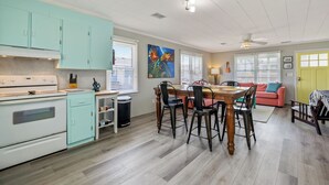 A kitchen with a stove, cabinets, and a dining area with chairs and a table. - The Way Back: Cozy Cottage Steps to Beach + Hot Tub + Coffee with Private Suite (Kill Devil Hills)