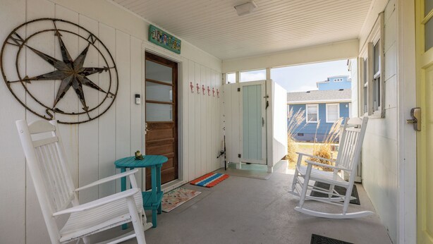 A porch with white rocking chairs, a wooden door, and a blue table. - The Way Back: Cozy Cottage Steps to Beach + Hot Tub + Coffee with Private Suite (Kill Devil Hills)