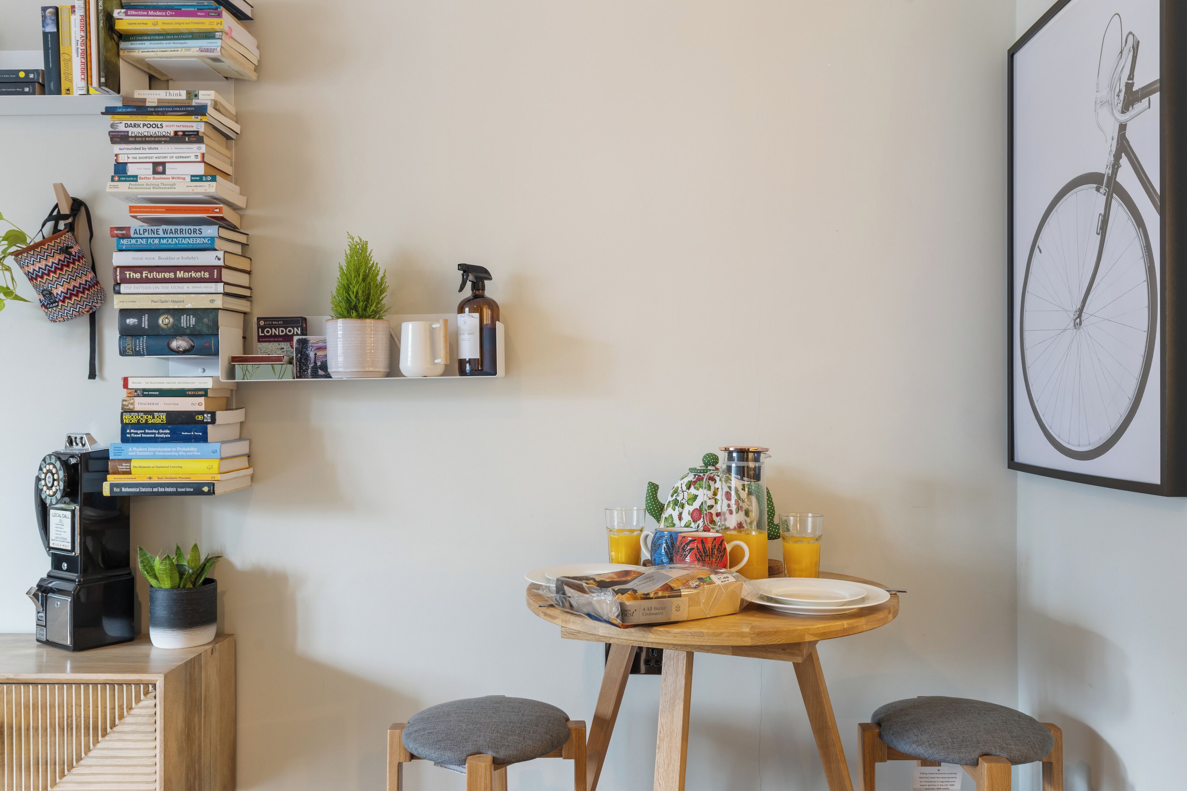 Une pièce chaleureuse avec une table en bois, deux chaises, une bibliothèque, une plante en pot et, au mur, un tableau représentant un vélo.