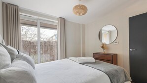 Un dormitorio moderno con una cama grande, una mesita de noche con un jarrón, un espejo redondo y una puerta oscura.