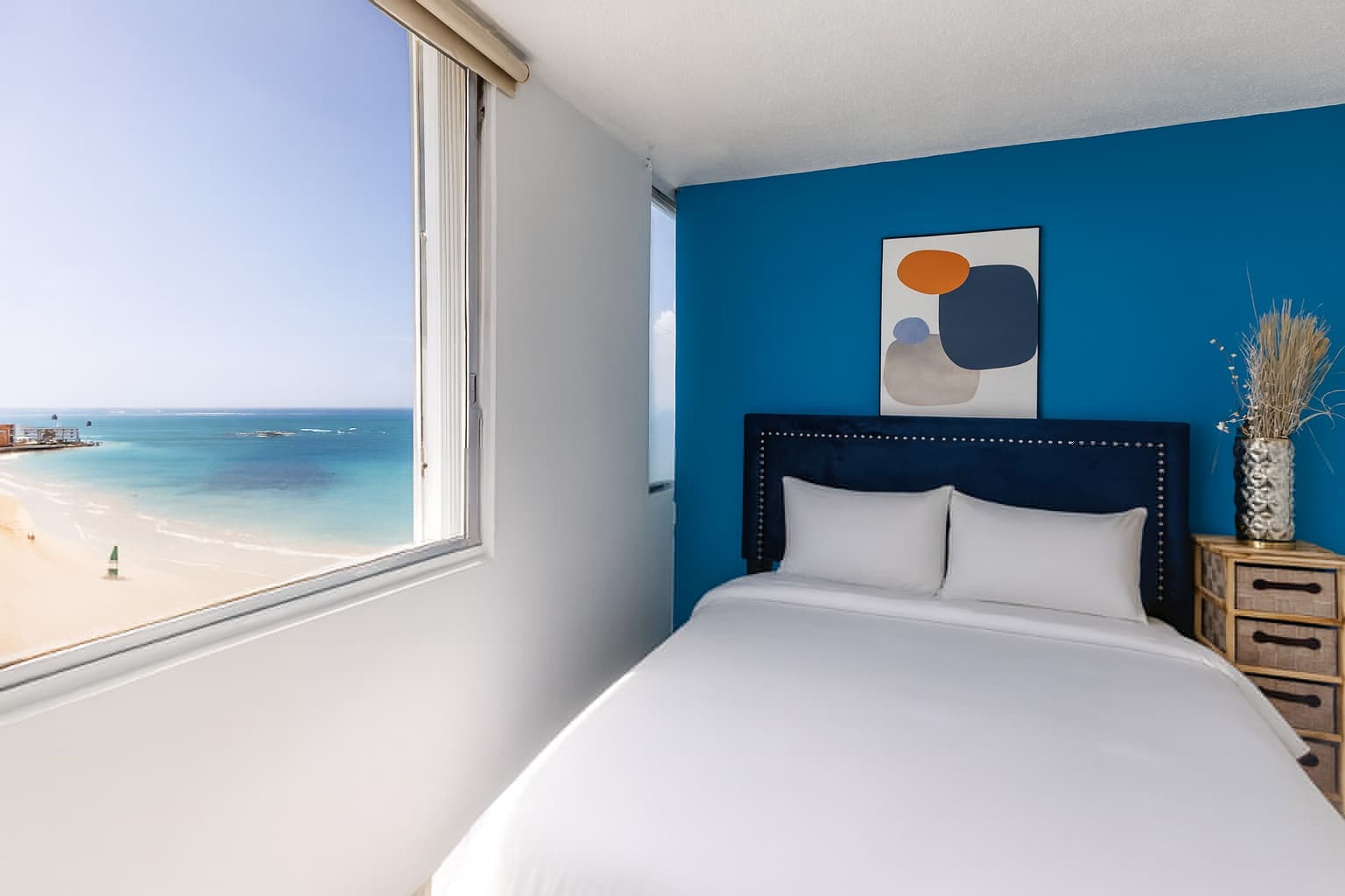 Un dormitorio con un ventanal grande con vistas a una playa, una cama con ropa blanca, una pared azul decorativa y un adorno sobre la cama.