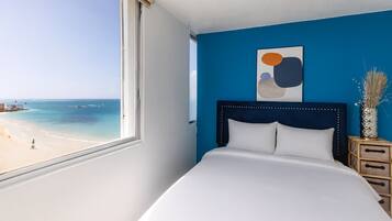 Un dormitorio con un ventanal grande con vistas a una playa, una cama con ropa blanca, una pared azul decorativa y un adorno sobre la cama.