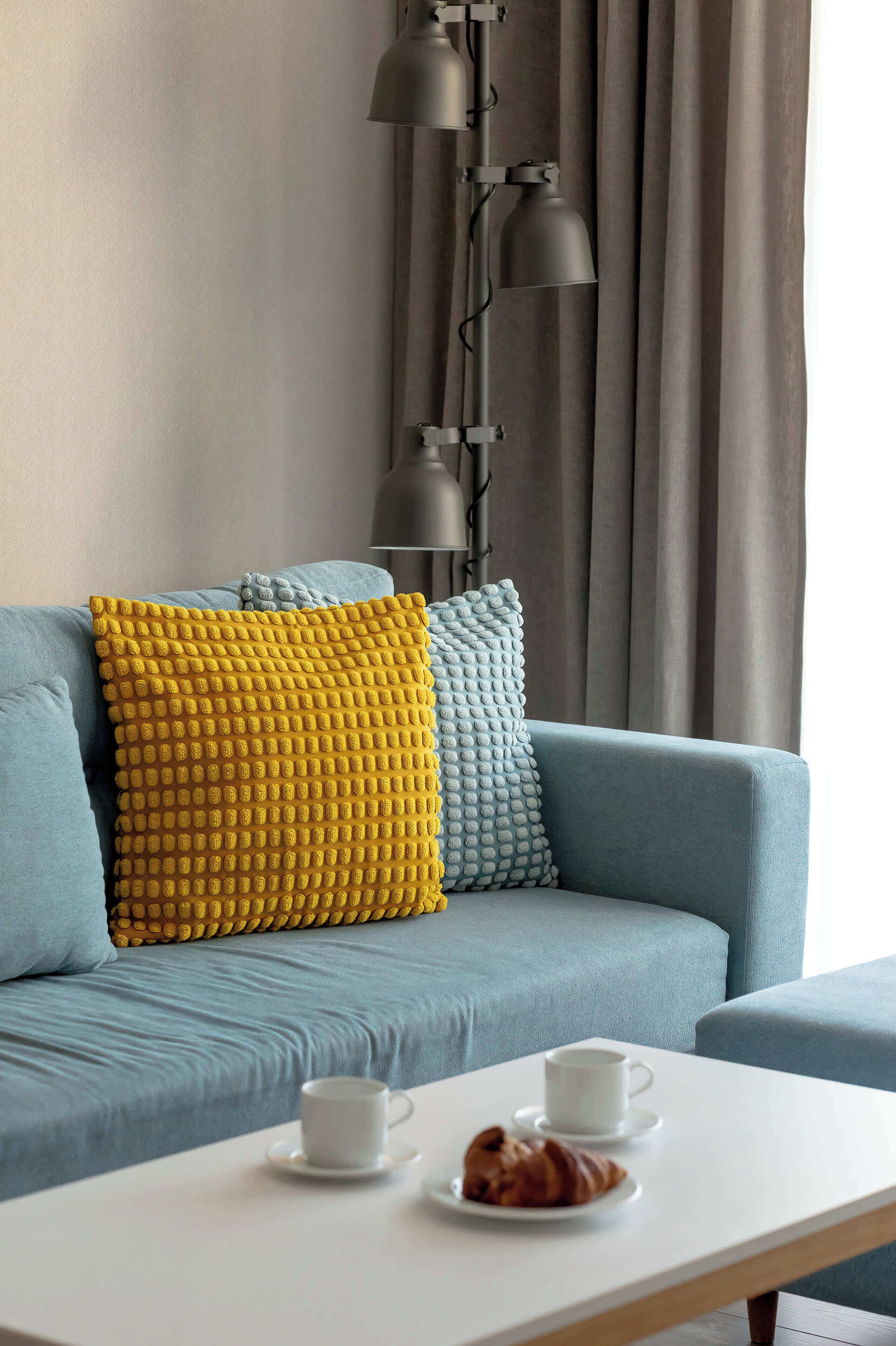 Una sala de estar moderna con un sofá azul, dos cojines estampados, una mesa blanca con una taza y un croissant, y una lámpara de pie.