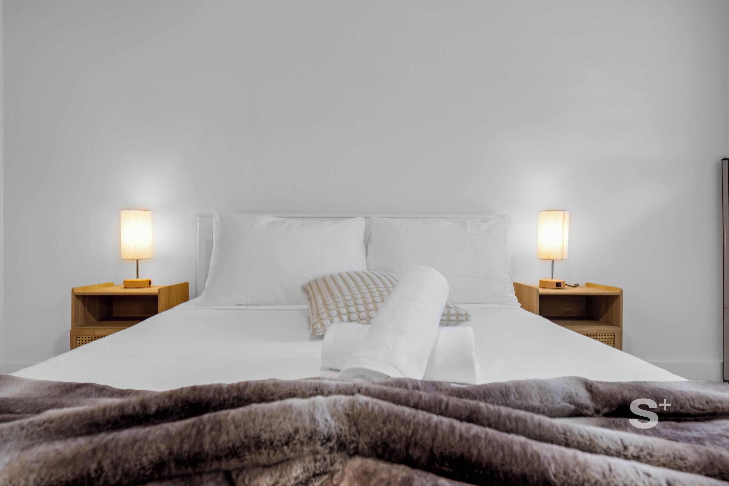 A neatly made bed with white linens, two bedside lamps, and a dark fur throw blanket.