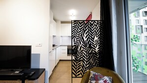 Ein modernes Wohnzimmer mit Fernseher, einer Couch mit Zierkissen und Blick auf einen Balkon mit Grünpflanzen.