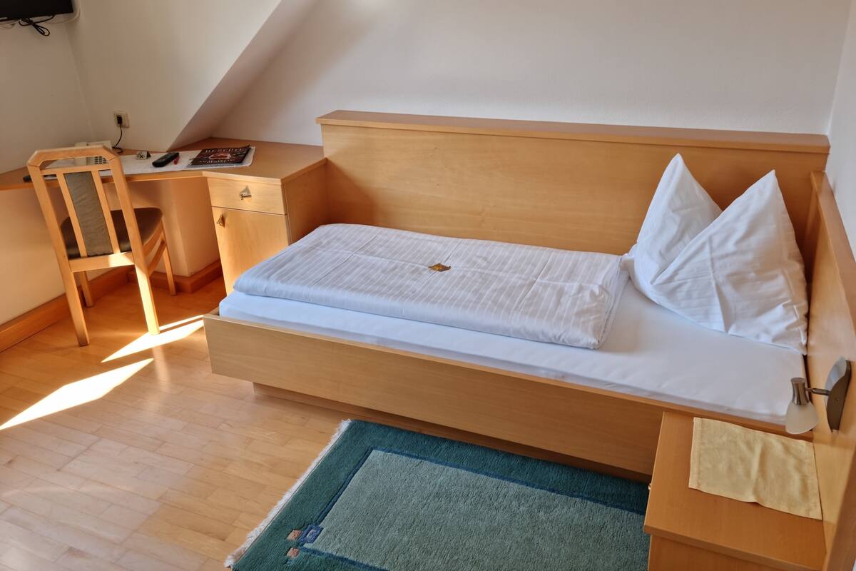 A single bed with a wooden headboard, a bedside table, a chair, and a desk with a chair.