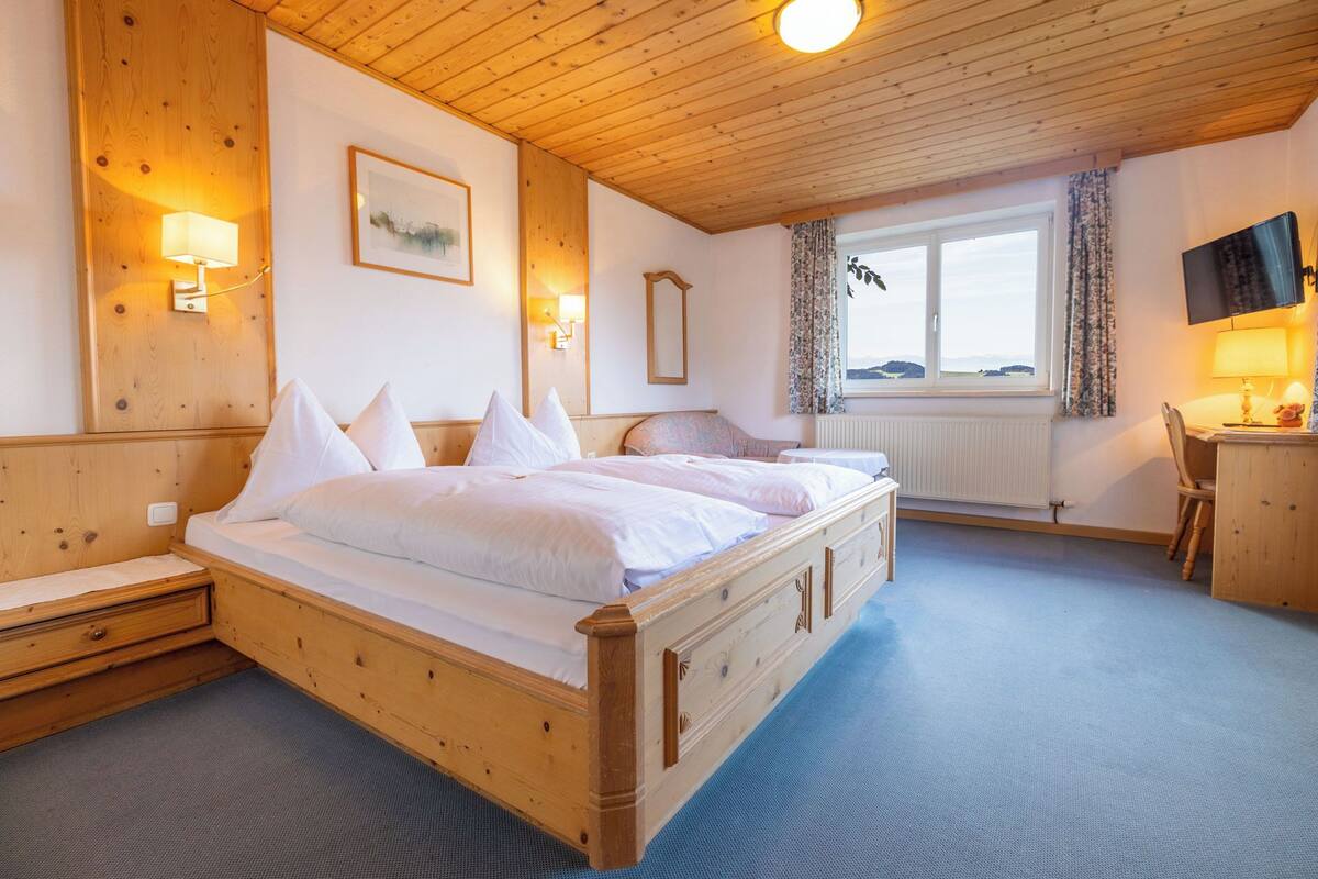 A wooden four-poster bed with white linens, a small bedside table, a desk with a chair, a window with a view, and a mounted TV.