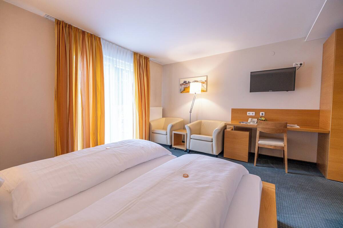 A hotel room with a bed, two chairs, a desk, a lamp, a TV, and a window with curtains.