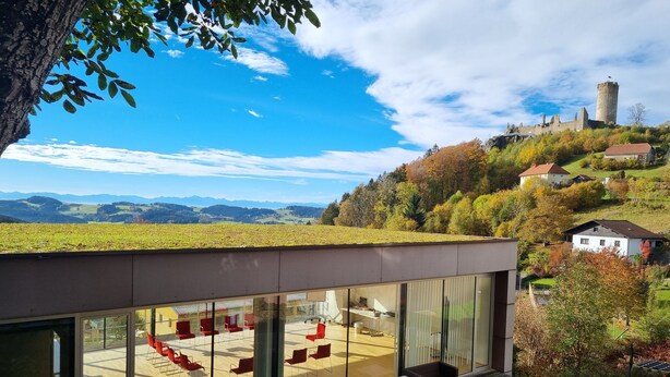A modern building with large glass windows, outdoor seating, and a view of a castle and hills. - Landhotel Hoftaverne Atzmüller (Oberneukirchen)