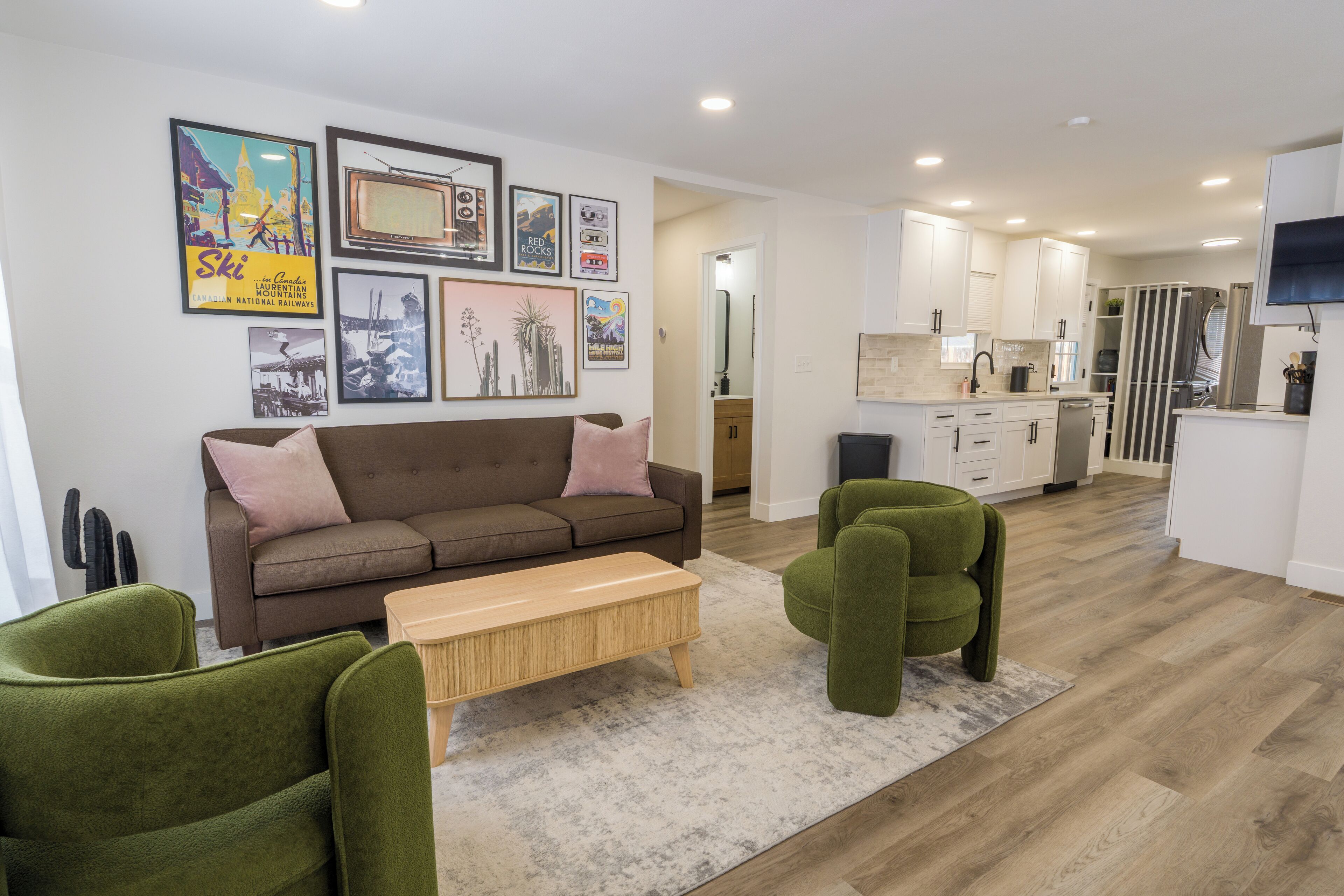 A modern living room with a brown sofa, green armchairs, and a wooden coffee table. A kitchen with white cabinets and a stainless steel refrigerator is visible in the background.