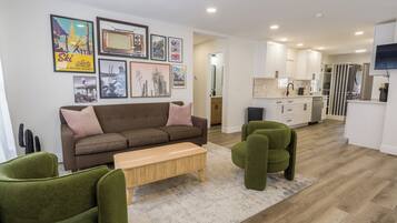 A modern living room with a brown sofa, green armchairs, and a wooden coffee table. A kitchen with white cabinets and a stainless steel refrigerator is visible in the background.