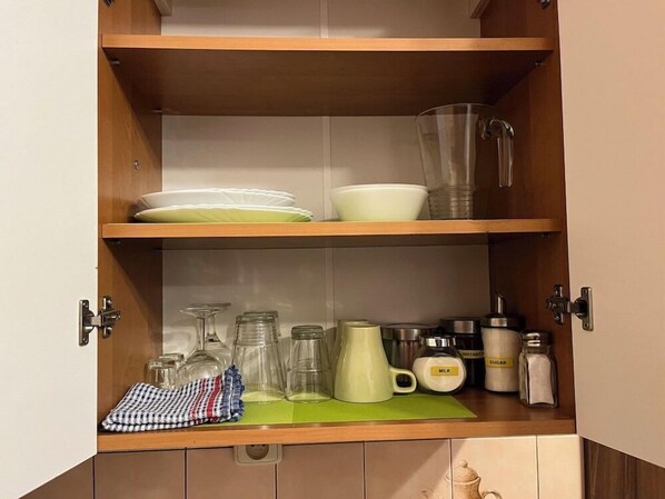 A kitchen cabinet with shelves holding dishes, glasses, a kettle, and kitchen essentials.