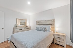 Une chambre à coucher avec un grand lit, une tête de lit en bois, une table de chevet avec des tiroirs, une petite table de chevet avec une lampe et un miroir accroché au mur.