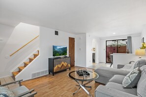 Un salon moderne avec un escalier en bois, un téléviseur à écran plat, un coin repas avec une table et des chaises, et une vue à travers une porte vitrée.
