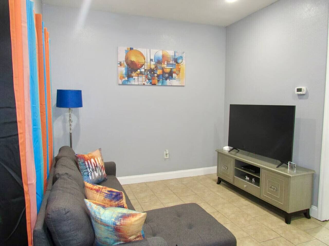 Ein Wohnzimmer mit grauem Sofa, bunten Kissen, einem Flachbildfernseher auf einem grauen Schrank und einem an der Wand hängenden abstrakten Kunstwerk.