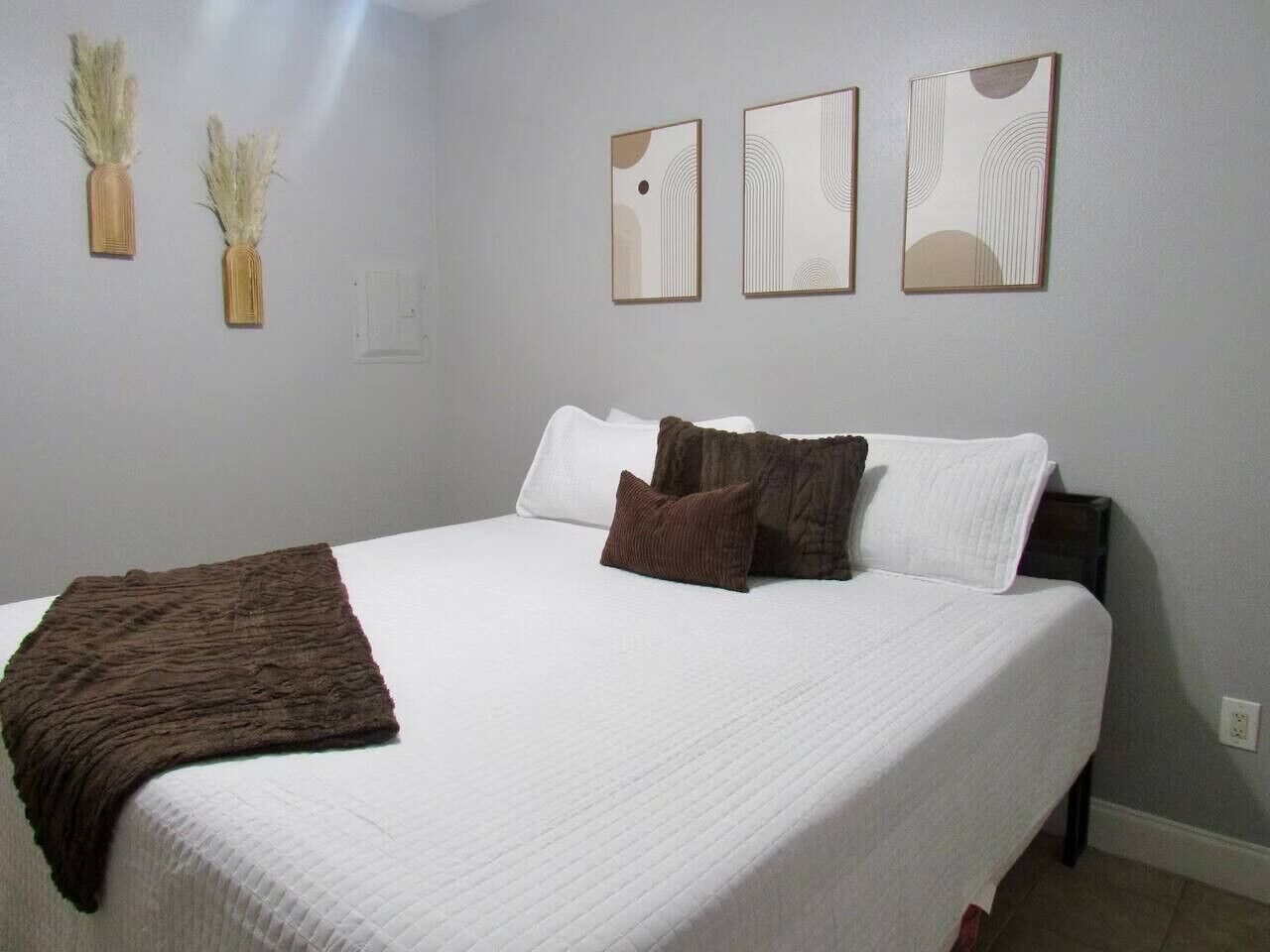Una cama bien tendida con ropa de cama blanca y marrón, una manta marrón y tres cuadros abstractos en la pared.