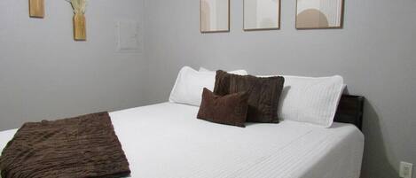 Una cama bien tendida con ropa de cama blanca y marrón, una manta marrón y tres cuadros abstractos en la pared.