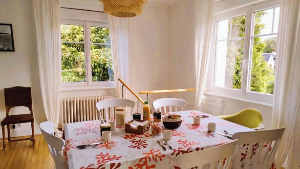 Un comedor con una mesa dispuesta para cuatro, sillas blancas, una lámpara de techo tejida y ventanas con follaje visible.