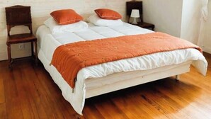 Un dormitorio con techo inclinado, una cama con ropa de cama blanca y naranja, una silla de madera, una mesita de noche y una lámpara colgante.