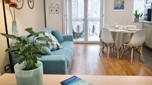 Ein modernes Wohnzimmer mit einer Couch, einem Esstisch mit Stühlen, einer Topfpflanze und einem Buch mit dem Titel 'SARDINIA' auf dem Tisch.