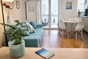 A modern living room with a sofa, a dining table with chairs, a potted plant, and a book titled 'SARDINIA' on the table. - Cozy bright apartment with balcony for three people, pets allowed (Cagliari)