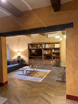 A spacious living room with a herringbone-patterned wooden floor, a dark sofa, a coffee table, and a glass dining table. There is a bookshelf filled with books and decorative items, and a television on a wooden stand. - Cozy 2-bedroom apartment in the historic center of Anghiari (Anghiari)