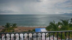 A beach view from a balcony with a railing, palm trees, and a distant mountain range. - Beachfront One Beach Street Condo - Pool - In the Heart of Old Town PV (Puerto Vallarta)