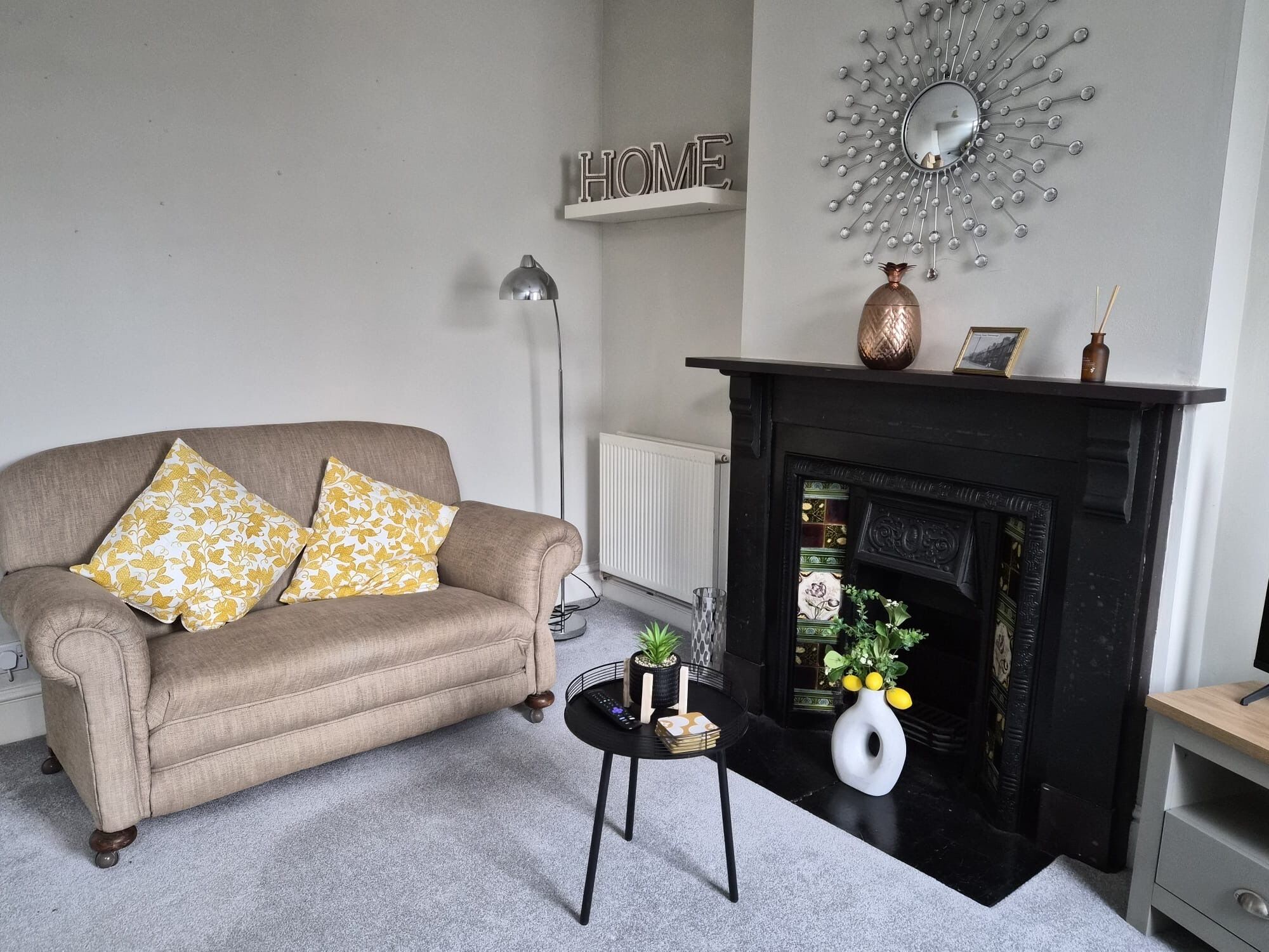 Un salon avec un canapé beige, deux coussins à motifs, une lampe sur pied, une petite table d’appoint avec un vase et un cadre photo, et une cheminée agrémentée d’objets décoratifs.