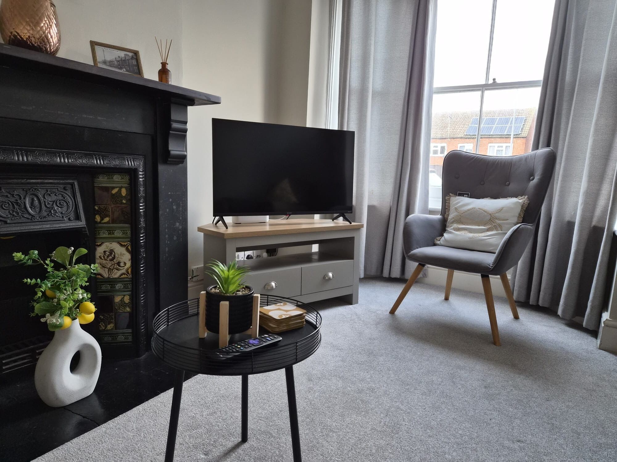 Ein Wohnzimmer mit Kamin, einem Flachbildfernseher auf einem Holzboden, einem grauen Sessel und einem kleinen runden Tisch mit Pflanze und Blumenvase.