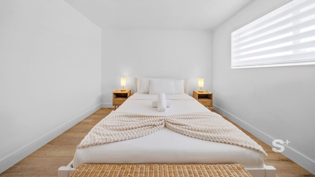Une chambre à coucher au style minimaliste, avec un grand lit, deux tables de chevet dotées de lampes et une fenêtre avec des stores.