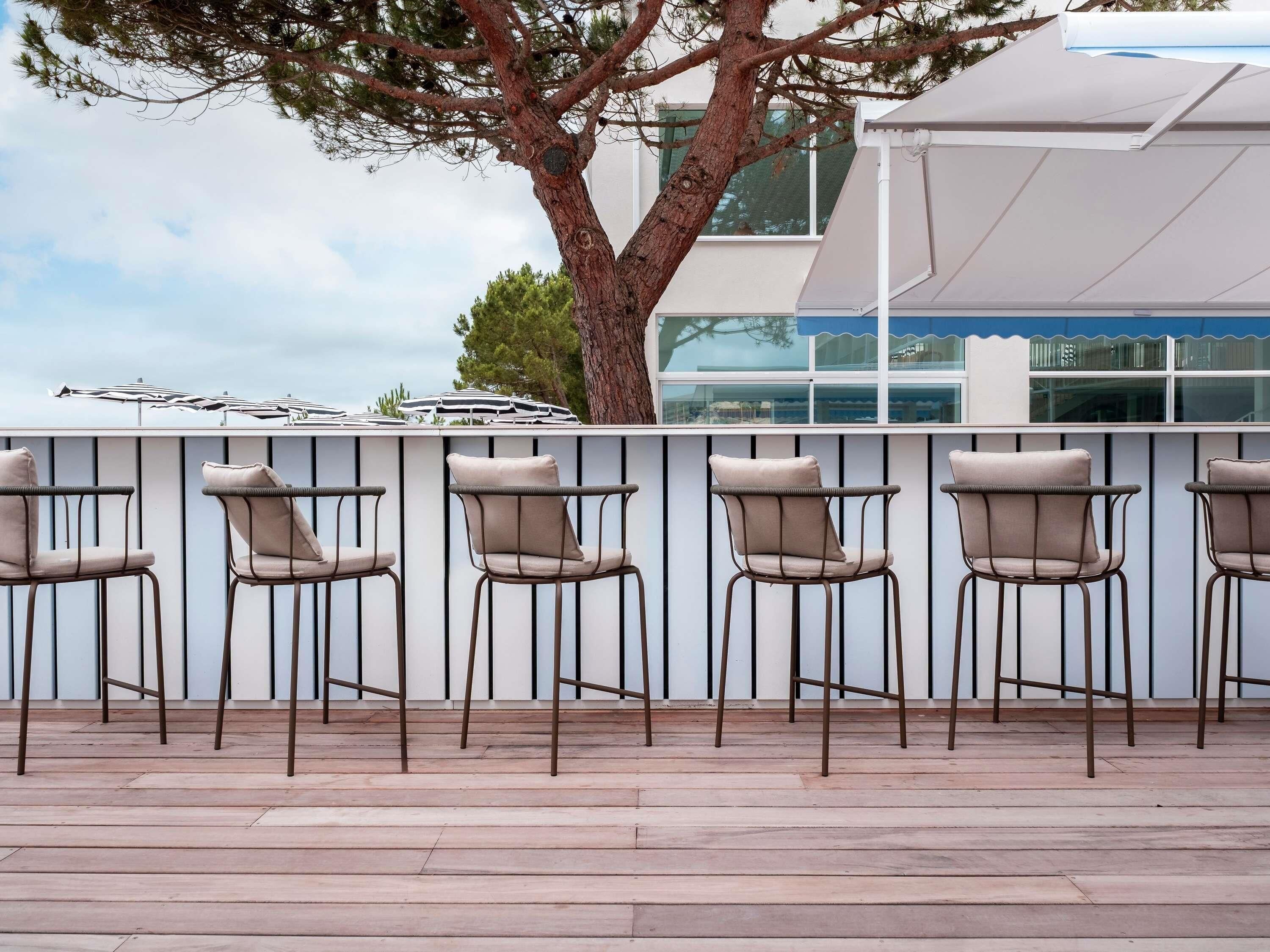 A row of outdoor bar-style chairs with metal frames and cushioned seats, aligned against a white wall with vertical lines.
