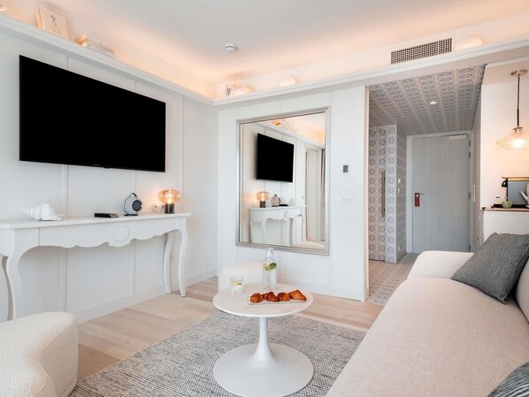 A modern living room with a white sofa, a round coffee table, a TV mounted on the wall, a console table with decorative items, and a mirror above a vanity. - Bel Hôtel Oléron Thalasso & Spa - Mgallery (Ouverture 2026) (Saint-Trojan-les-Bains)