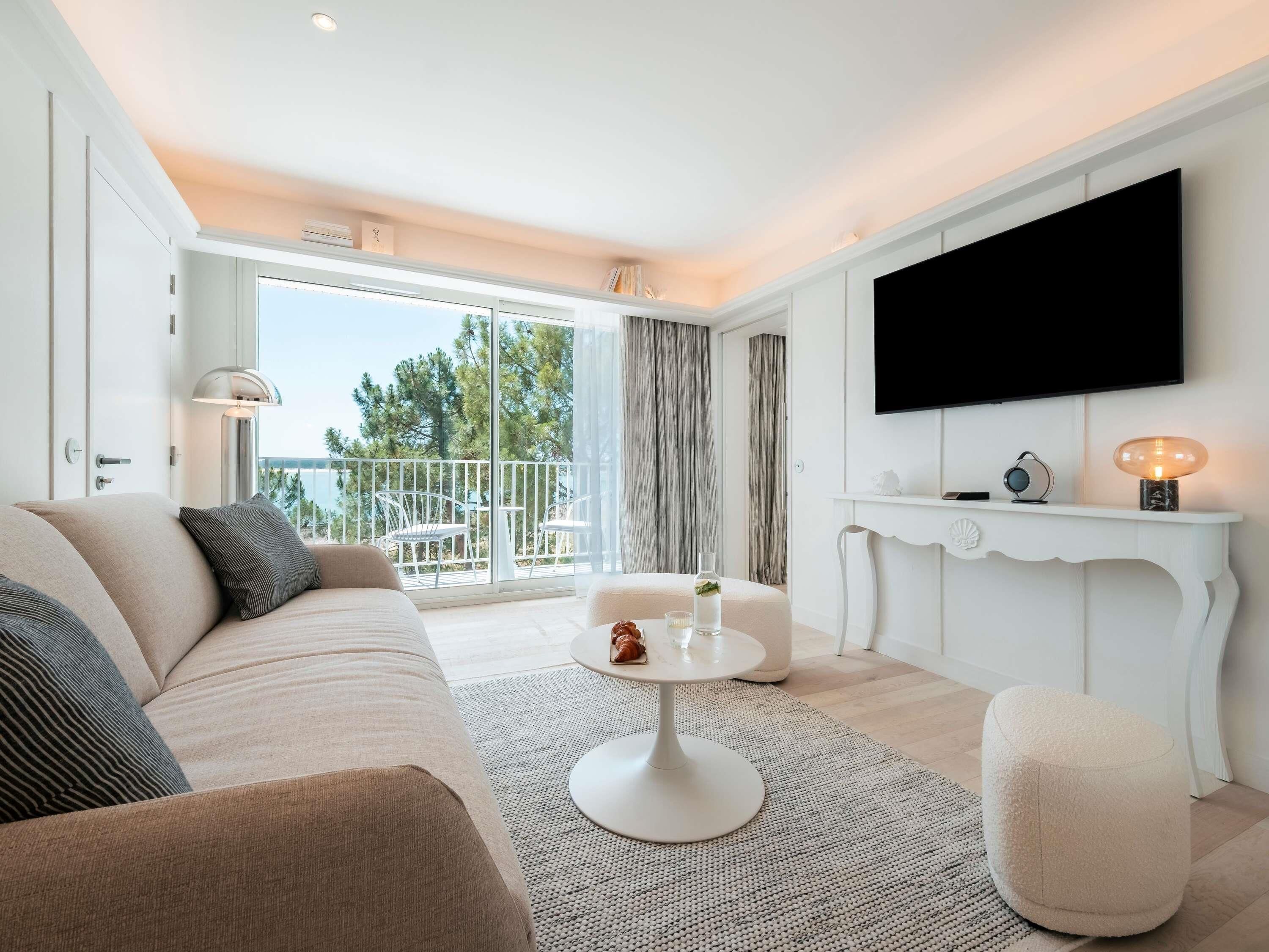 A modern living room with a sofa, a round coffee table, a white console table with a TV, and a balcony with outdoor furniture.