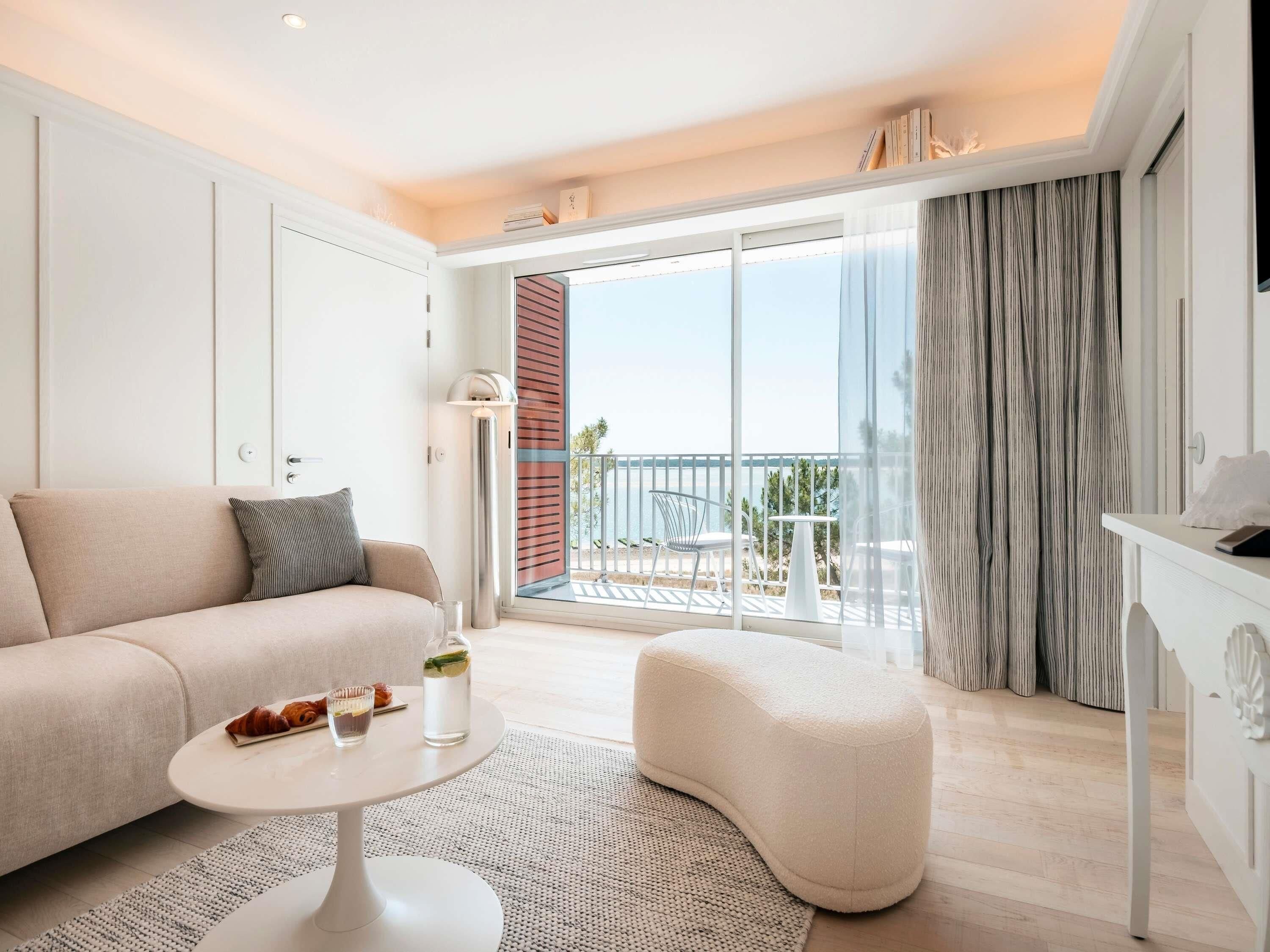 A modern hotel room with a sofa, a small round table, a pouf, a balcony with a view, and a large window.