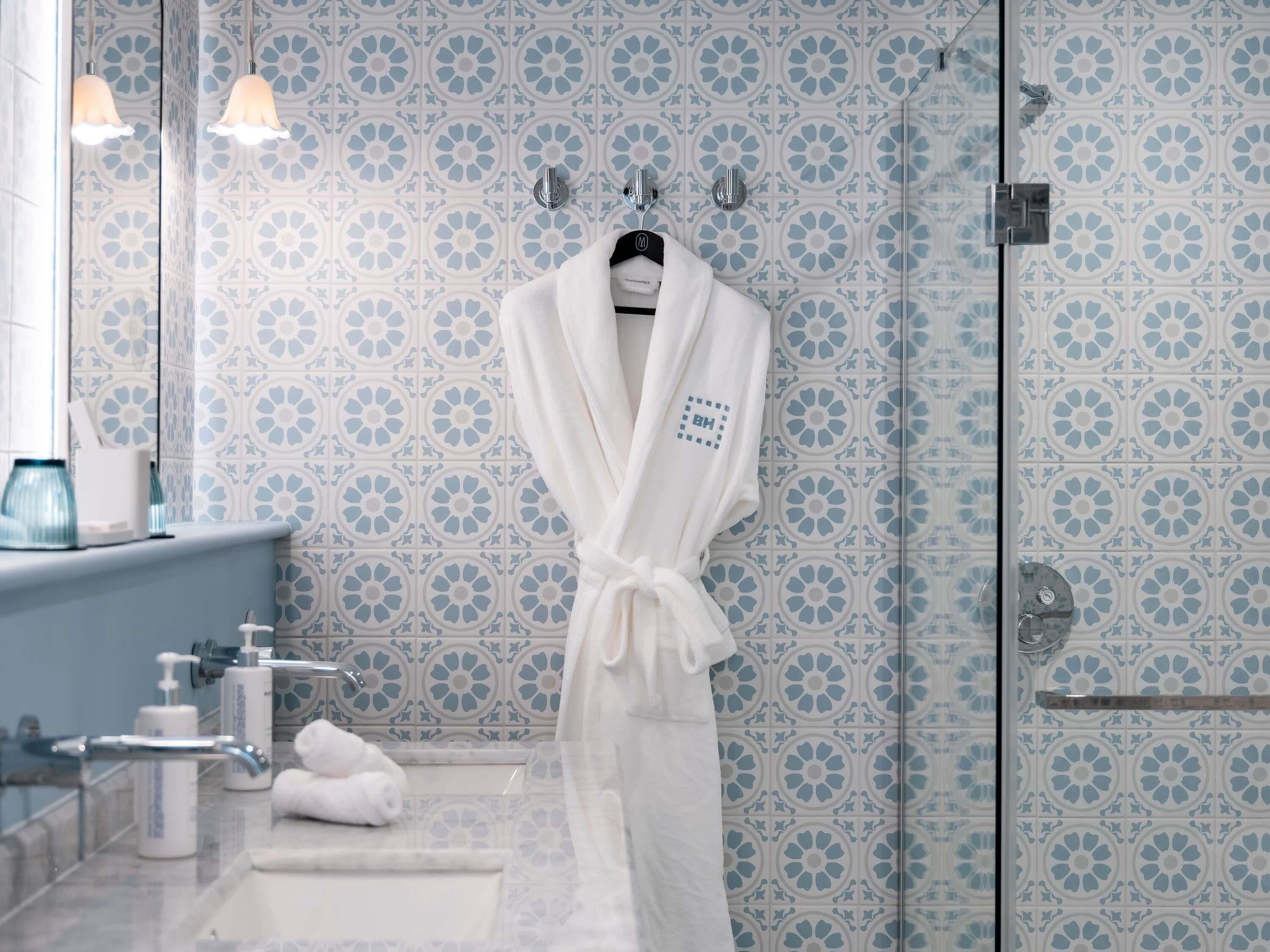 A bathroom with a patterned tile wall, a white robe hanging on a hook, a glass shower enclosure, and a sink with a towel on it.