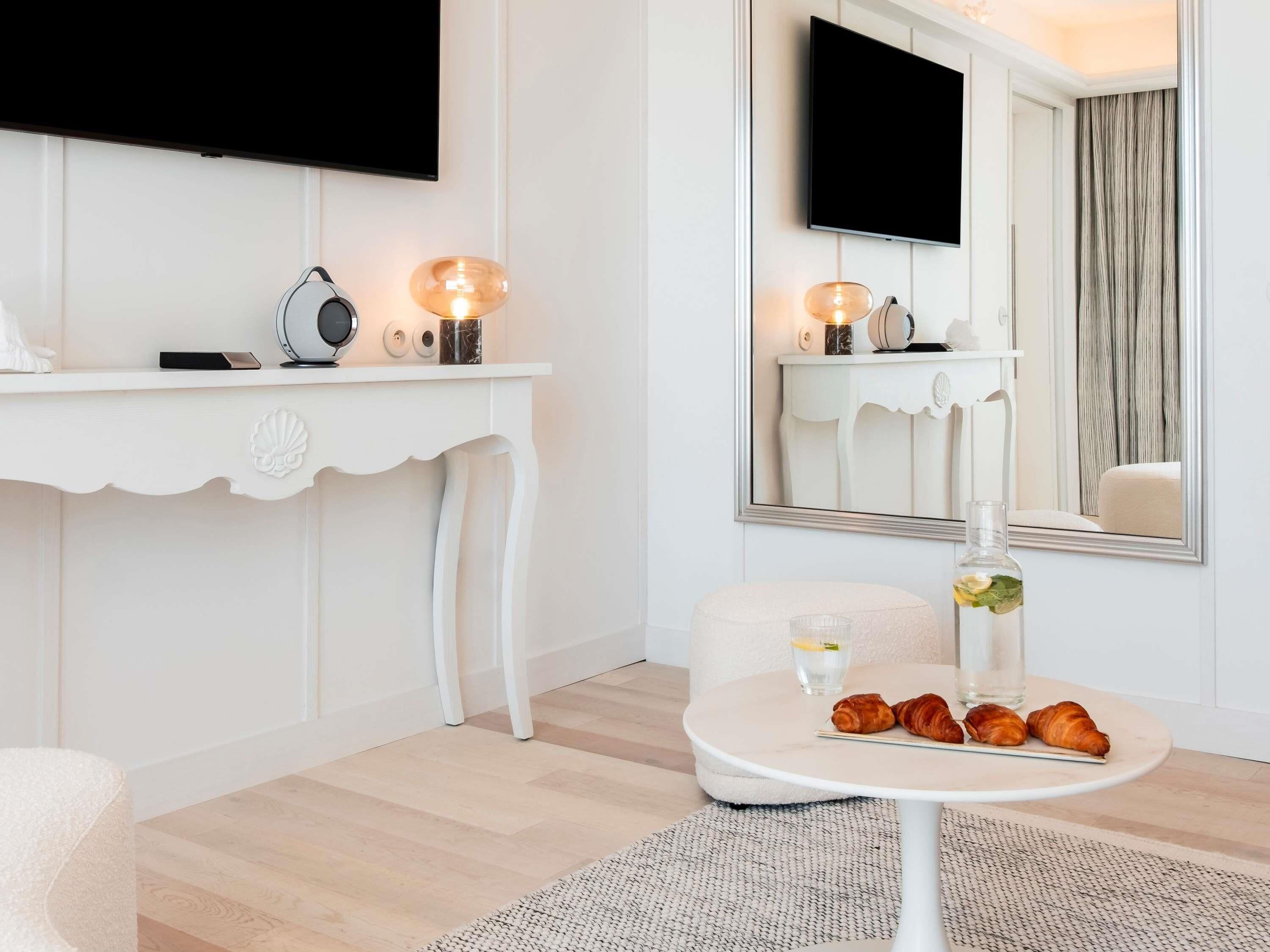 A modern hotel room with a white console table, a round table with croissants, a large mirror, and a flat-screen TV mounted on the wall.