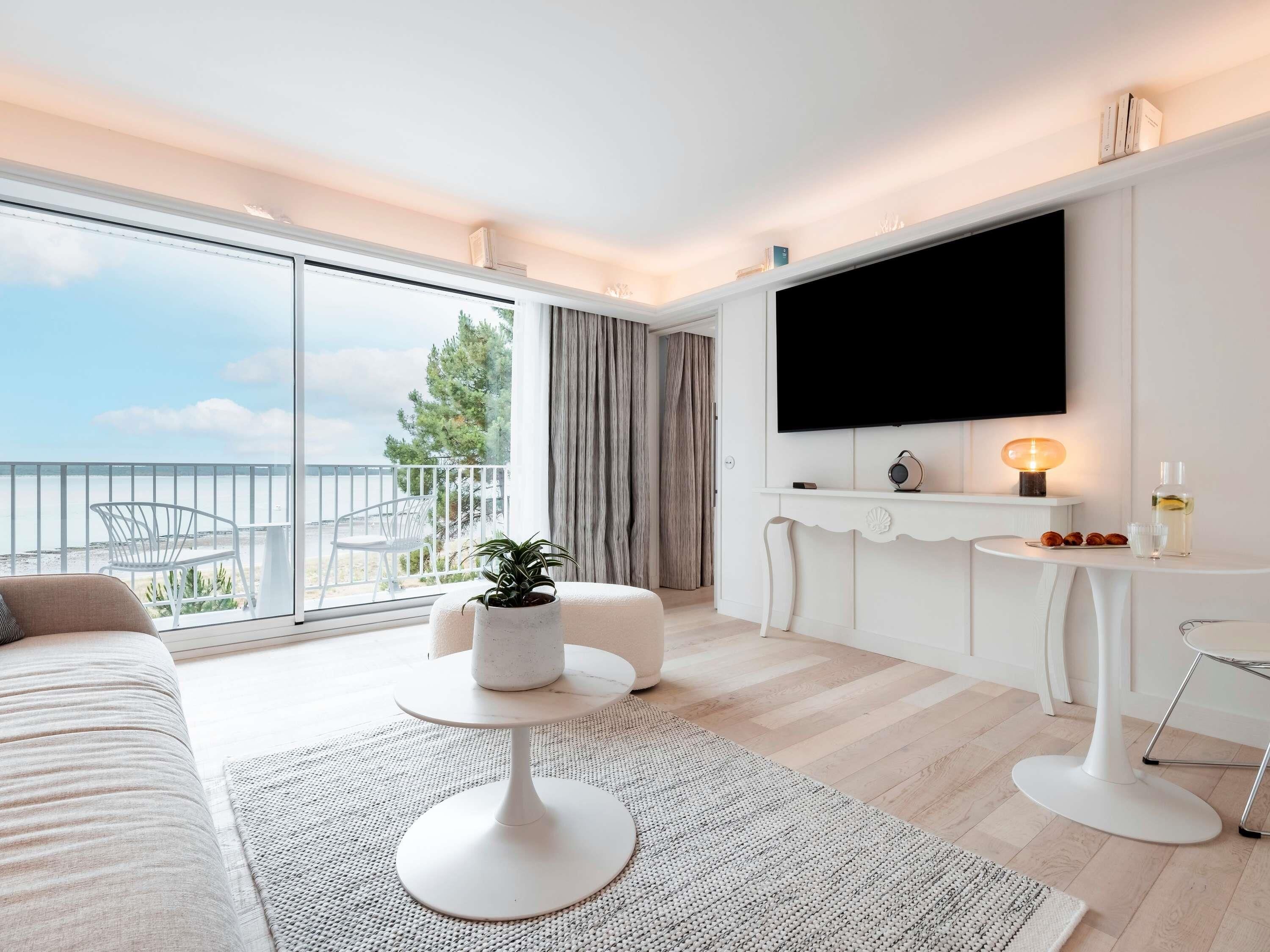 Een moderne hotelkamer met een ruim balkon, een flatscreen televisie, een tafeltje met lamp en een zithoek met een bank en voetenbank.