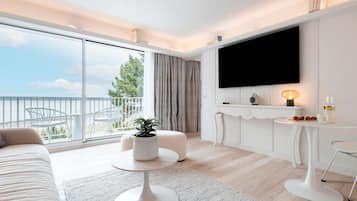 Een moderne hotelkamer met een ruim balkon, een flatscreen televisie, een tafeltje met lamp en een zithoek met een bank en voetenbank.