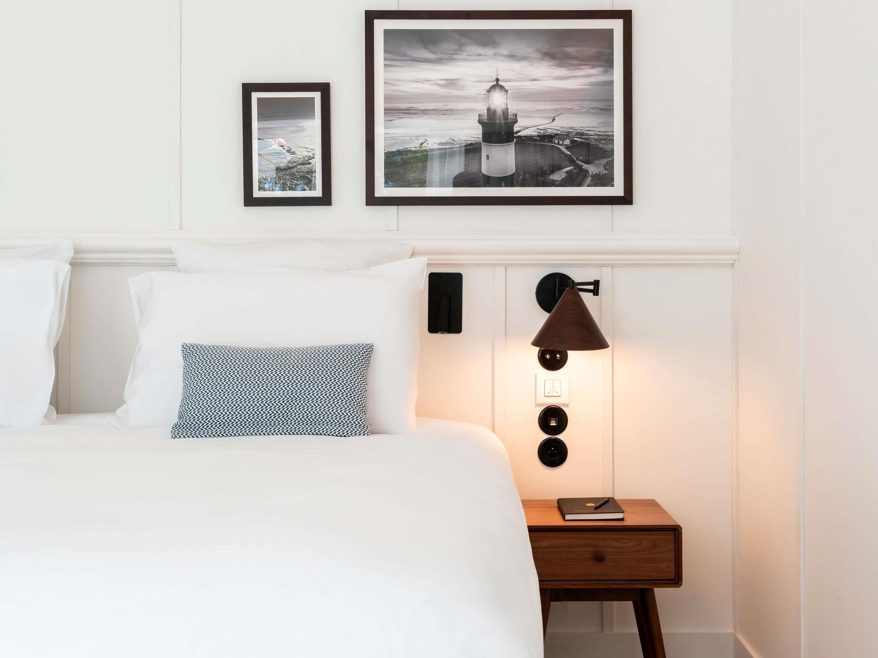 Una cama bien hecha con sábanas blancas y almohadas estampadas, una mesita de noche con lámpara, y dos cuadros en la pared.
